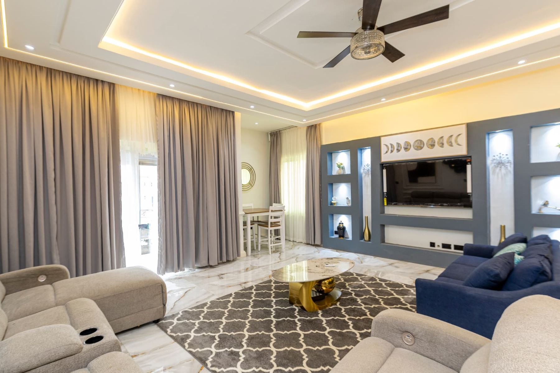 Unit 107 living room with recliner sofas, navy couch, ceiling fan and illuminated media wall